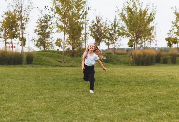 Une petite fille joyeuse aux cheveux longs traverse l'herbe du parc et se réjouit