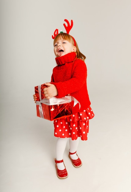 Petite fille joyeuse dans un pull rouge tient une boîte cadeau rouge sur fond blanc.