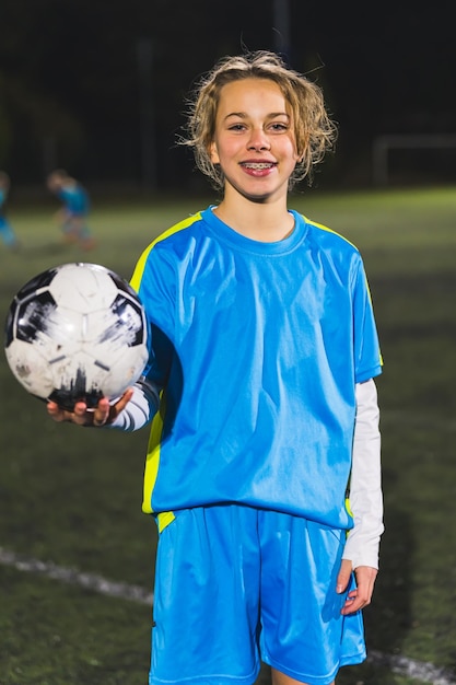 Une petite fille joyeuse et heureuse en uniforme bleu tenant un ballon de football et prenant une photo