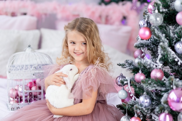 Petite fille avec lapin à l'arbre de Noël dans une belle robe rose