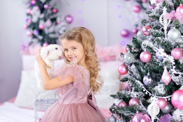 Petite fille avec lapin à l'arbre de Noël dans une belle robe rose