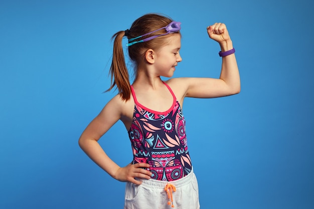 La petite fille lève le bras et regarde les muscles porte des lunettes et un maillot de bain