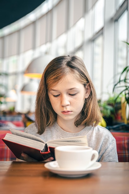 Une petite fille lit un livre assise dans un café avec une tasse de thé