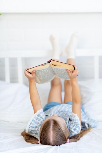 Une petite fille lit un livre sur le lit à la maison sur un lit en coton blanc allongé sur le dos