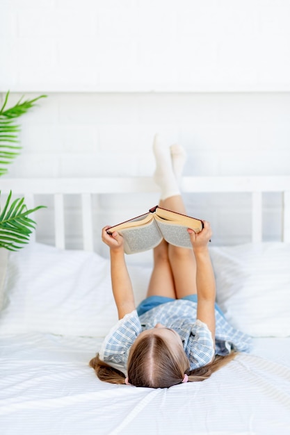 Une petite fille lit un livre sur le lit à la maison sur un lit en coton blanc allongé sur le dos