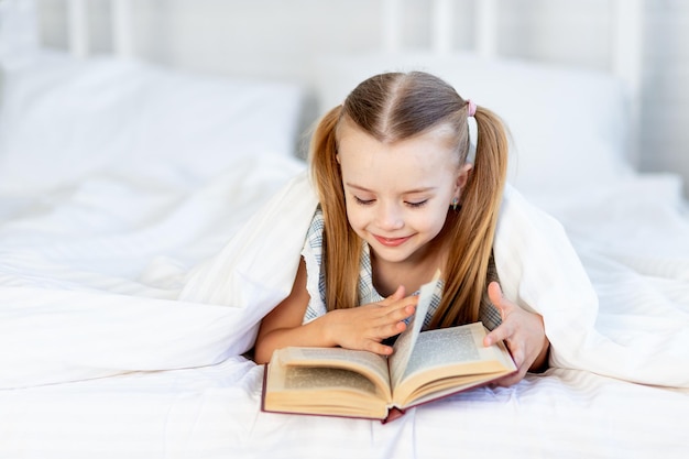 Une petite fille lit un livre sur le lit à la maison sur un lit en coton blanc sous une couverture et sourit doucement