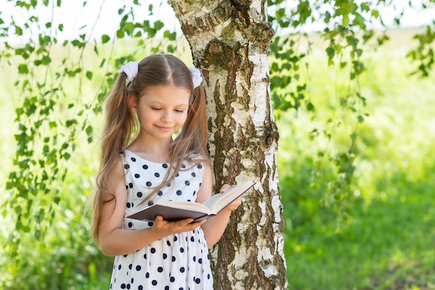 Une petite fille lit un livre par une journée d'été ensoleillée sous un arbre.