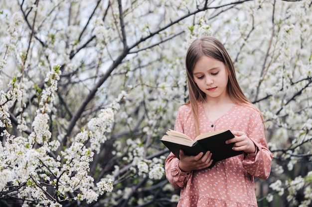 Petite fille lit un livre près d'un arbre en fleurs dans le jardin.