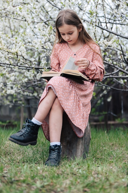 Petite fille lit un livre près d'un arbre en fleurs dans le jardin.
