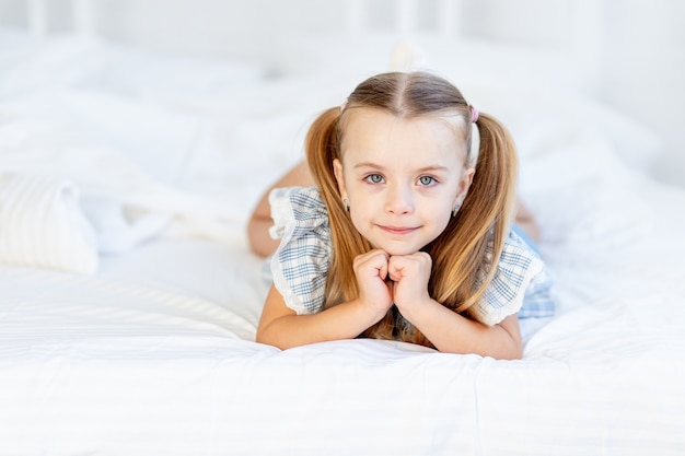Une petite fille sur le lit à la maison sur un lit en coton blanc se trouve et sourit doucement.