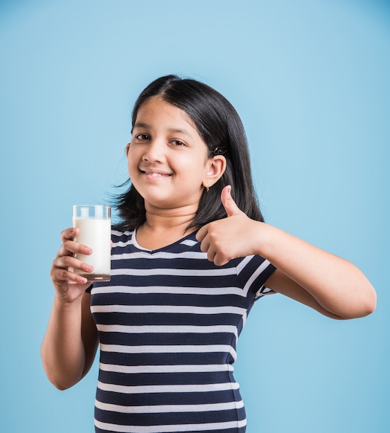 Petite fille ludique indienne ou asiatique mignonne tenant ou buvant un verre plein de lait, isolée sur fond coloré