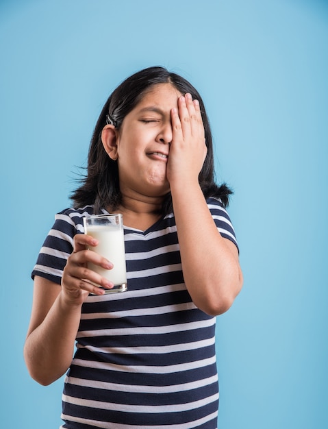 Petite fille ludique indienne ou asiatique mignonne tenant ou buvant un verre plein de lait, isolée sur fond coloré