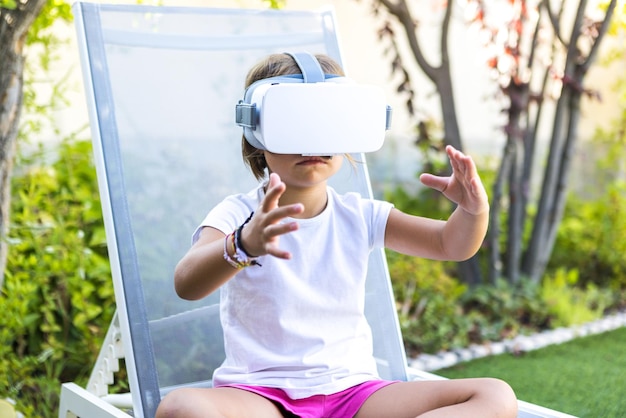 Petite fille avec des lunettes de réalité virtuelle essayant de toucher quelque chose de manière virtuelle assise sur une chaise longue dans le jardin de sa maison Concept numérique et de simulation de jeu Metaverse VR