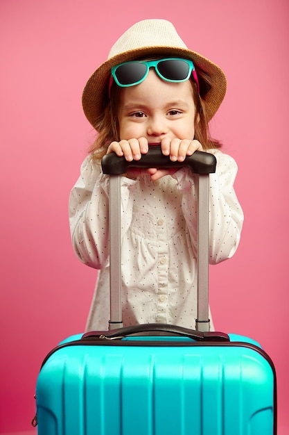 Petite fille à lunettes de soleil et chapeau de paille tenant une valise debout sur fond rose isolé