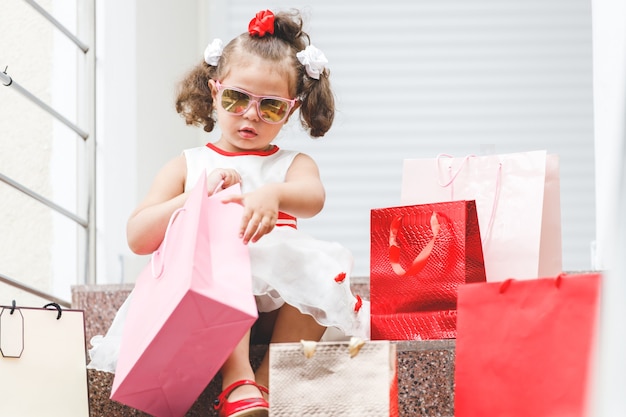 Une petite fille à lunettes de soleil est assise sur les marches du centre commercial avec des sacs colorés