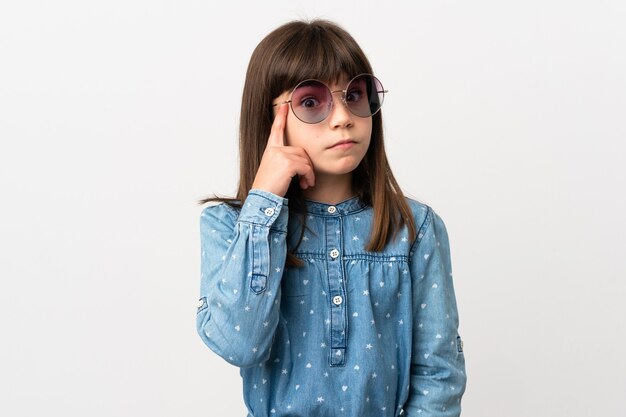 Petite fille avec des lunettes de soleil isolé sur fond blanc pensant une idée