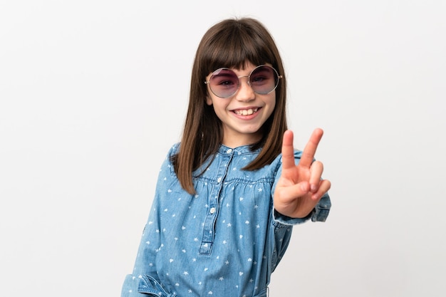 Petite fille avec des lunettes de soleil isolé sur un mur blanc souriant et montrant le signe de la victoire