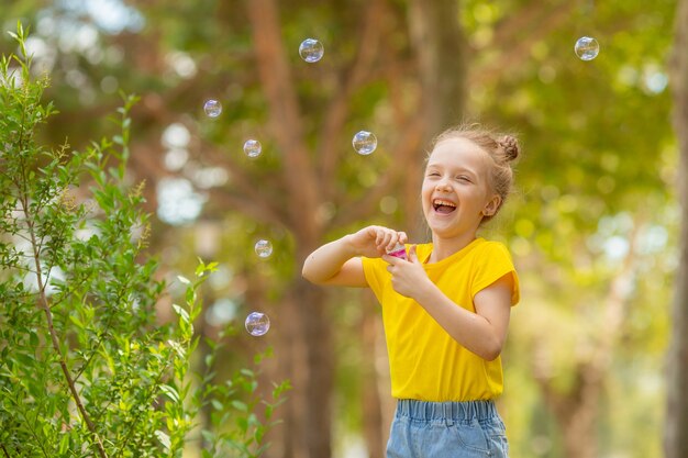 Une petite fille à lunettes de soleil souffle des bulles de savon dans le parc en été