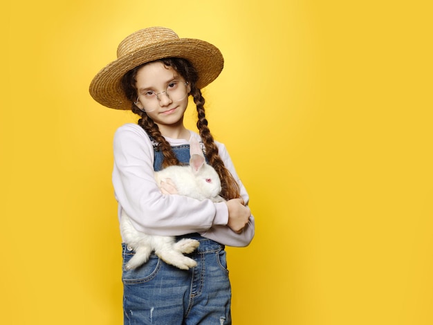 Petite fille à lunettes vêtue d'un jean et d'un chapeau de paille tenant un vrai lapin blanc isolé sur fond jaune