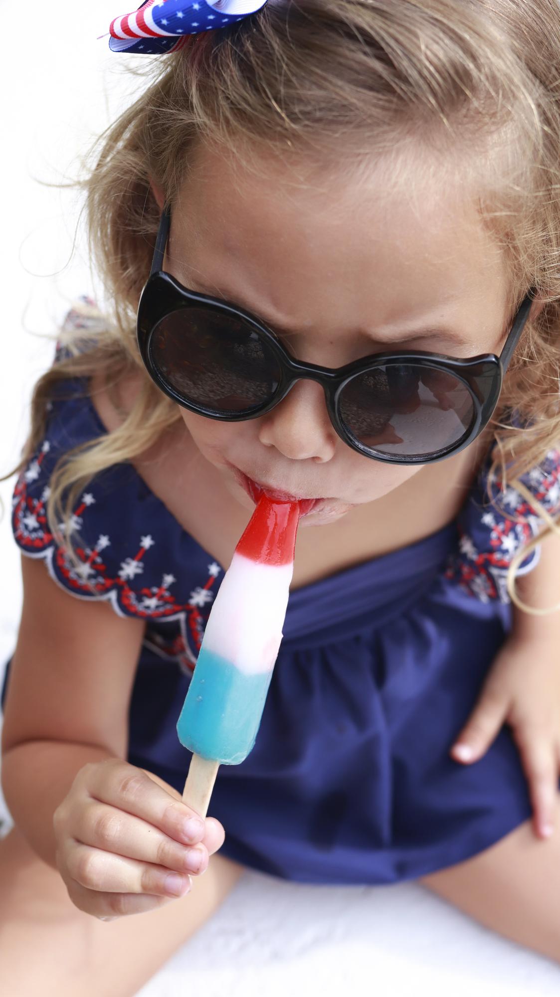 Une petite fille en maillot de bain bleu mangeant une glace en rouge, blanc et bleu.