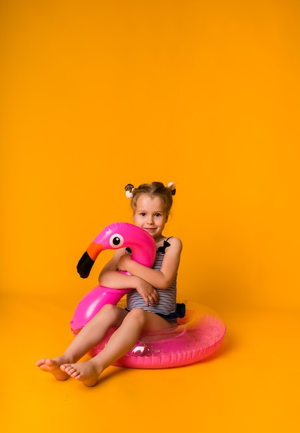 Petite fille en maillot de bain à rayures est assise sur un cercle gonflable rose avec un flamant rose sur une surface jaune avec un espace pour le texte