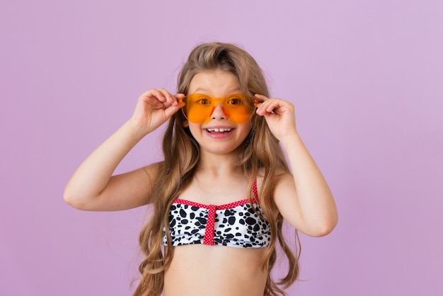 Une petite fille en maillot de bain sourit et s'accroche à ses lunettes orange.