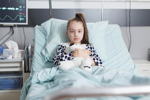 Petite fille malade tenant un ours en peluche et portant un tube à oxygène tout en se reposant seule dans son lit d'hôpital. Enfant malade dans la salle de récupération des patients de la clinique pédiatrique de soins de santé tout en regardant la caméra.