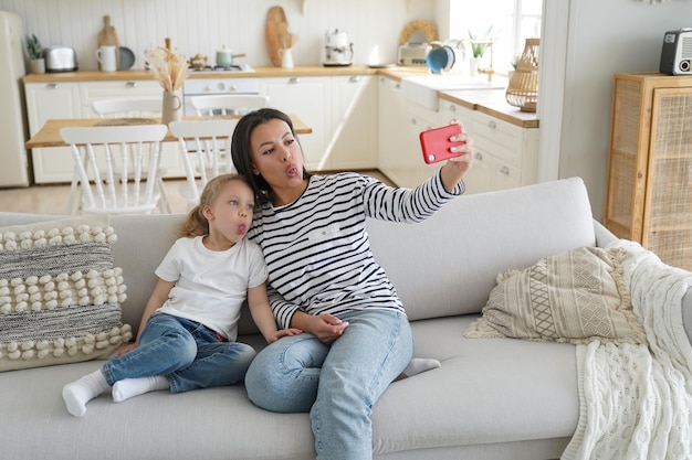 Petite fille maman montre des langues prendre selfie photo par smartphone assis sur un canapé ensemble à la maison