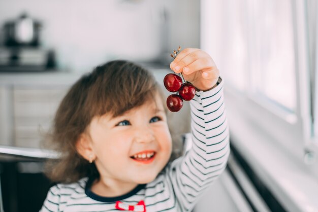 Une petite fille mange des cerises, tenant des baies dans ses mains et souriant mignon