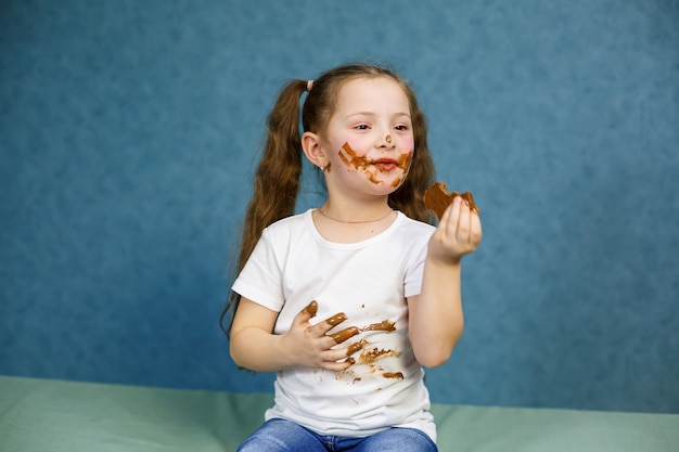 La petite fille mange du chocolat et tache son t-shirt blanc, son visage et lui tend les mains