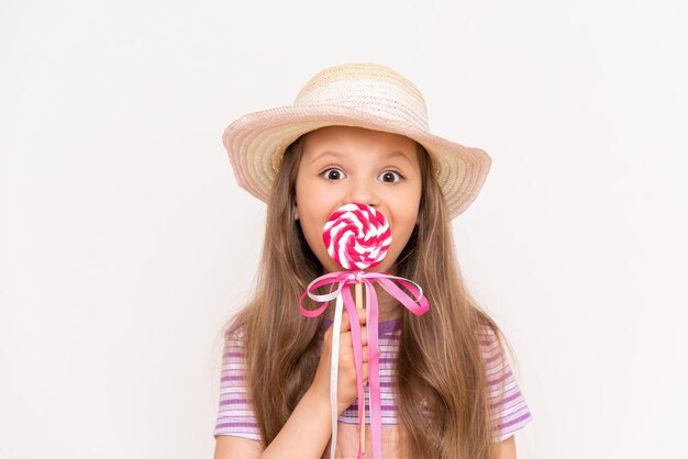 Une petite fille mange une sucette sur un fond blanc isolé Un enfant mange des bonbons dans un chapeau d'été en paille