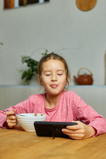 Petite fille mangeant des céréales avec du lait et regardant une vidéo sur un smartphone savoureuse nourriture saine Ralenti d'un bel enfant prenant son petit déjeuner à la maison dépendance du gadget