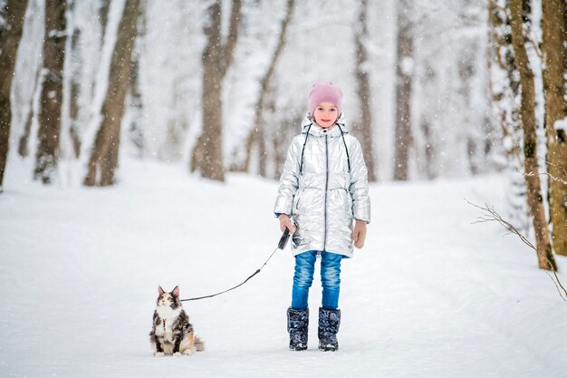 Une petite fille marche avec un chat en laisse dans un parc d'hiver enneigé