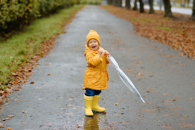 Une petite fille marche avec un parapluie dans des bottes en caoutchouc jaunes et un imperméable imperméable Autumn Walk