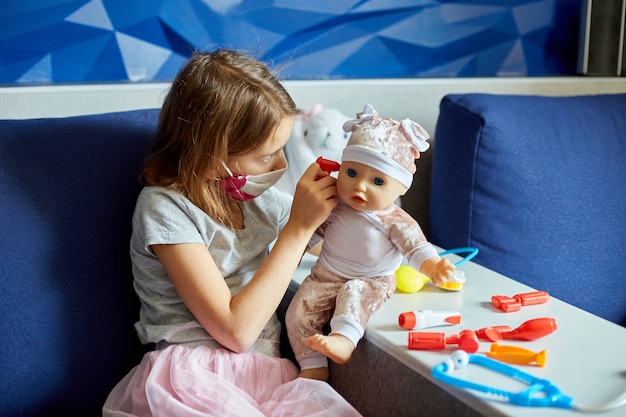 Une petite fille en masque médical est assise sur le canapé, joue un médecin, traite et écoute la poupée avec un stéthoscope dans un masque médical, à la maison