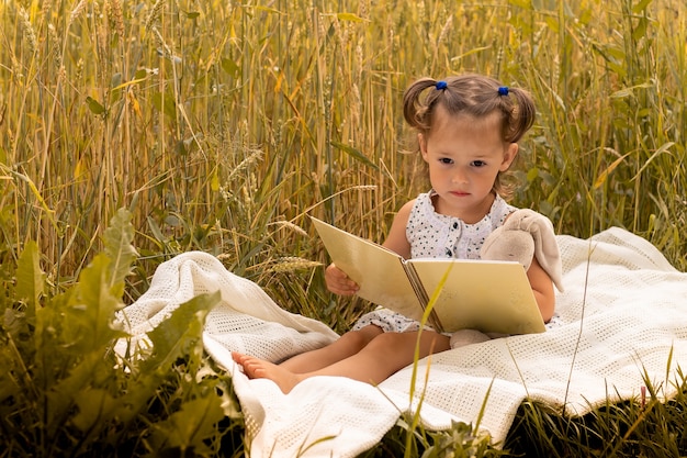 Petite fille mignonne 1-3 dans une robe légère embrasse un lièvre en peluche et lit un livre, assise sur un plaid dans un champ d'épillets de seigle en été.