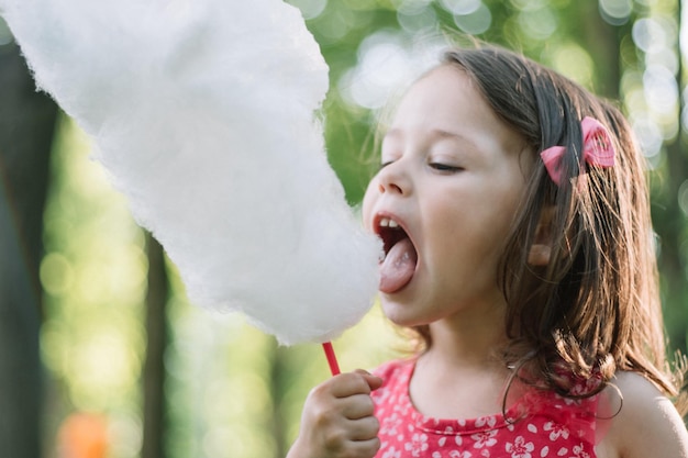 Petite fille mignonne 3-4 mangeant de la barbe à papa dans un parc ensoleillé, parmi de grands arbres sur l'herbe verte