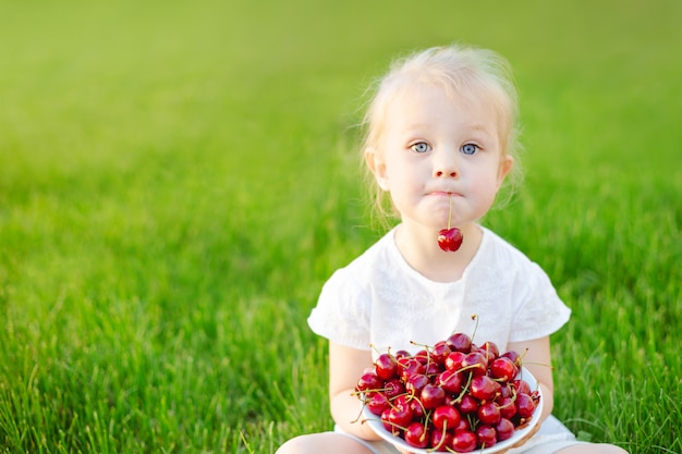 Une petite fille mignonne assise sur l&#39;herbe avec une assiette de cerises sur ses genoux et tenant une baie dans ses dents.