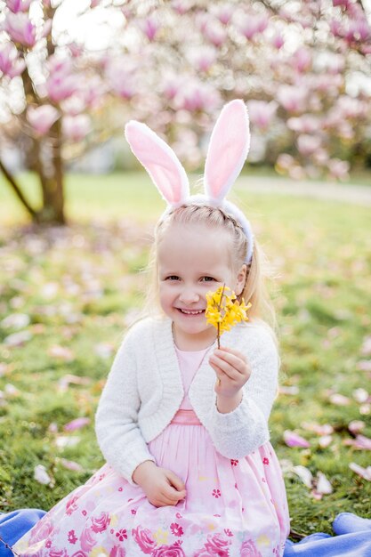 Petite fille mignonne assise sur l'herbe près du magnolia. Une fille habillée en lapin de Pâques tient une fleur et un œuf. Printemps.