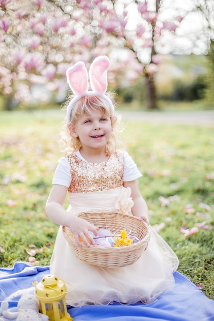 Petite fille mignonne assise sur l'herbe près du magnolia. Une fille habillée en lapin de Pâques tient une fleur et un œuf. Printemps.