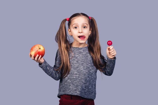 Petite fille mignonne avec un bonbon et une pomme dans ses mains. Choix de concept entre la nourriture saine et la malbouffe. isolé sur fond gris