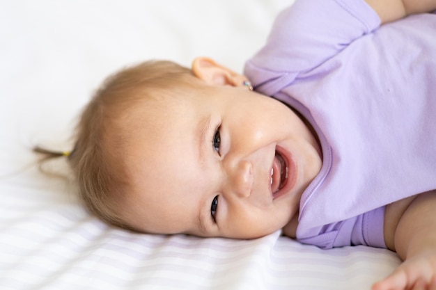 Petite fille mignonne en bonne santé dans un body est allongée sur le dos sur le lit sur une literie blanche, regarde la caméra, rit, gros plan. Le concept de produits pour bébés.