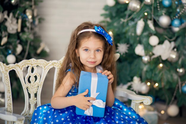 La petite fille mignonne avec un cadeau de Noël, dans une robe bleue s'assoit à côté des arbres de Noël décorés. Nouvel An, vacances en famille de Noël