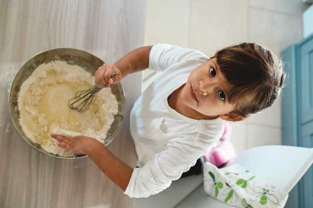 Une petite fille mignonne cuisine dans la cuisine tout en regardant la caméra ci-dessus S'amuser en faisant de la pizza ou de la pâte à pain Concept d'enfance et s'amuser en cuisinant