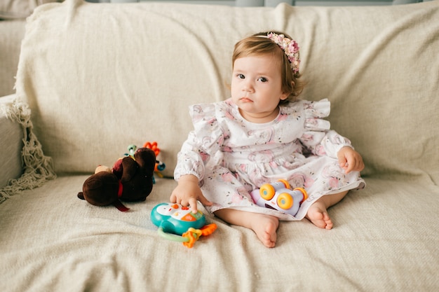 Petite fille mignonne dans une belle robe est assise sur le canapé et joue avec ses divers jouets.