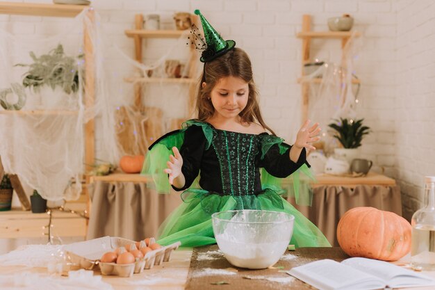 La petite fille mignonne dans un costume vert d'Halloween d'une sorcière ou d'une fée prépare une tarte à la citrouille