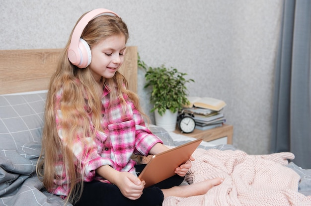 La petite fille mignonne dans des écouteurs utilise une tablette à la maison