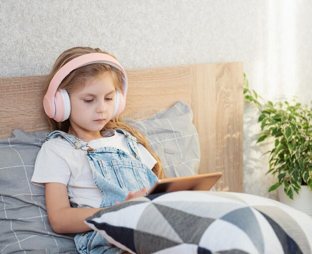 La petite fille mignonne dans des écouteurs utilise une tablette à la maison