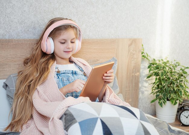 La petite fille mignonne dans des écouteurs utilise une tablette à la maison