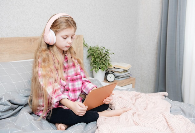 La petite fille mignonne dans des écouteurs utilise une tablette à la maison